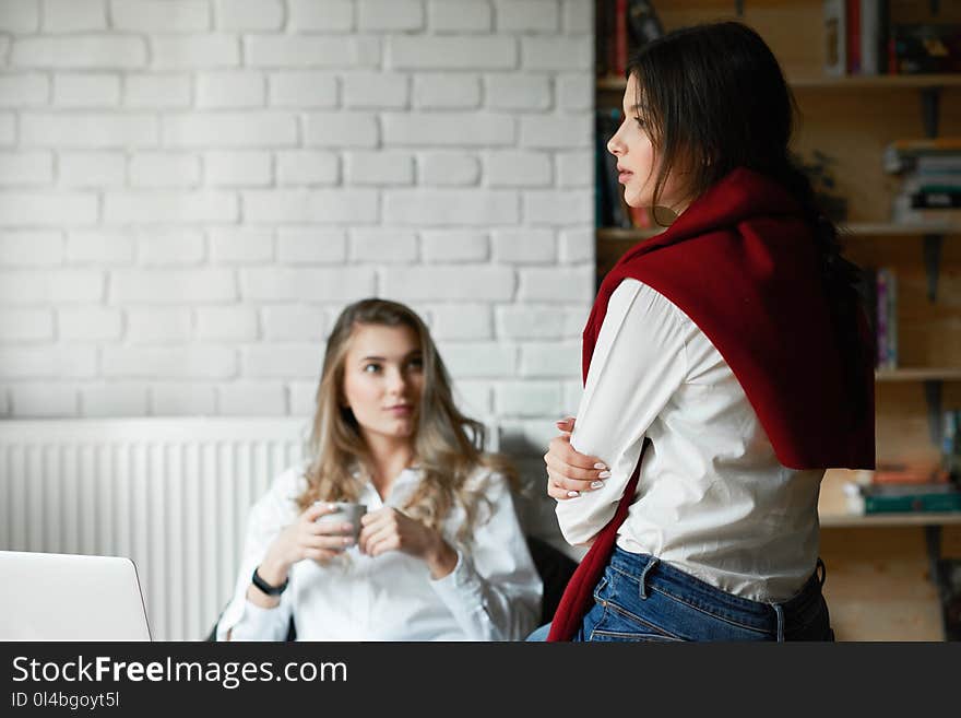 Two young female friends wearing casual classic white shirts communicating, working with laptop, sitting in spacy room with white walls. Drinking coffee, learning. Businesswomen, freelancers. Two young female friends wearing casual classic white shirts communicating, working with laptop, sitting in spacy room with white walls. Drinking coffee, learning. Businesswomen, freelancers.