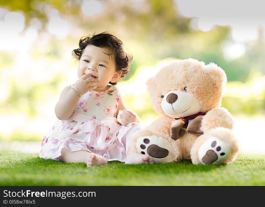 Baby Sitting on Green Grass Beside Bear Plush Toy at Daytime