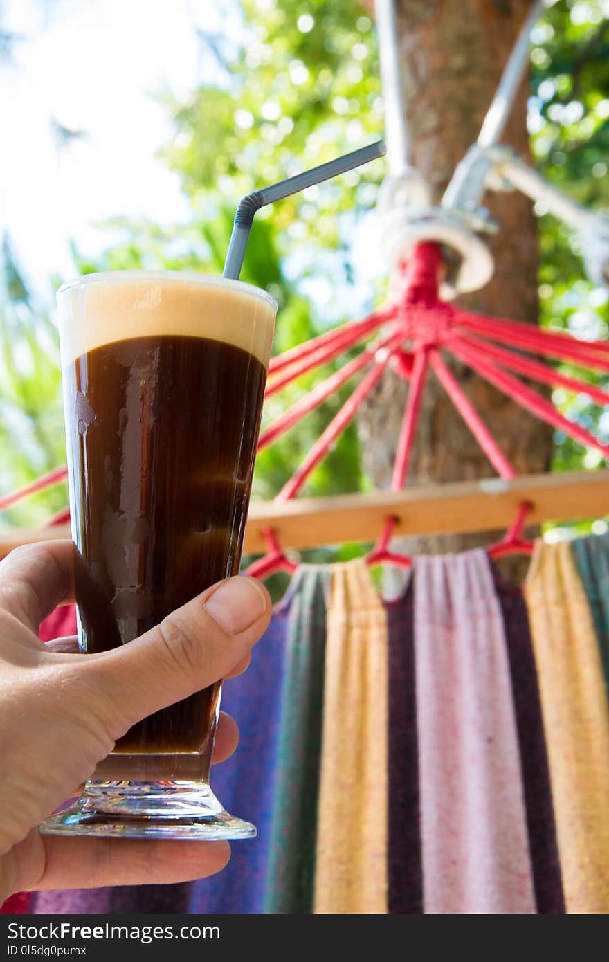 Close up of woman hand female lying on hammock holding glass of cold coffee. Refreshing/relaxing coffee time