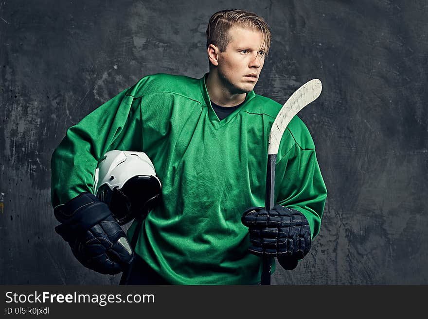 Tired professional hockey player in a green sportswear holds a hockey stick and protective helmet on a gray background.