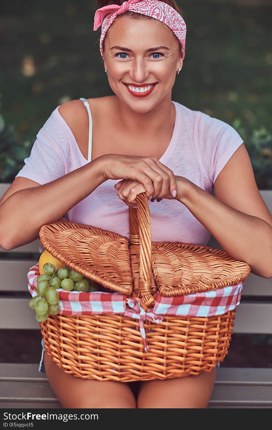 Happy beautiful redhead female wearing casual clothes holds a picnic basket while sitting on a bench in a park.