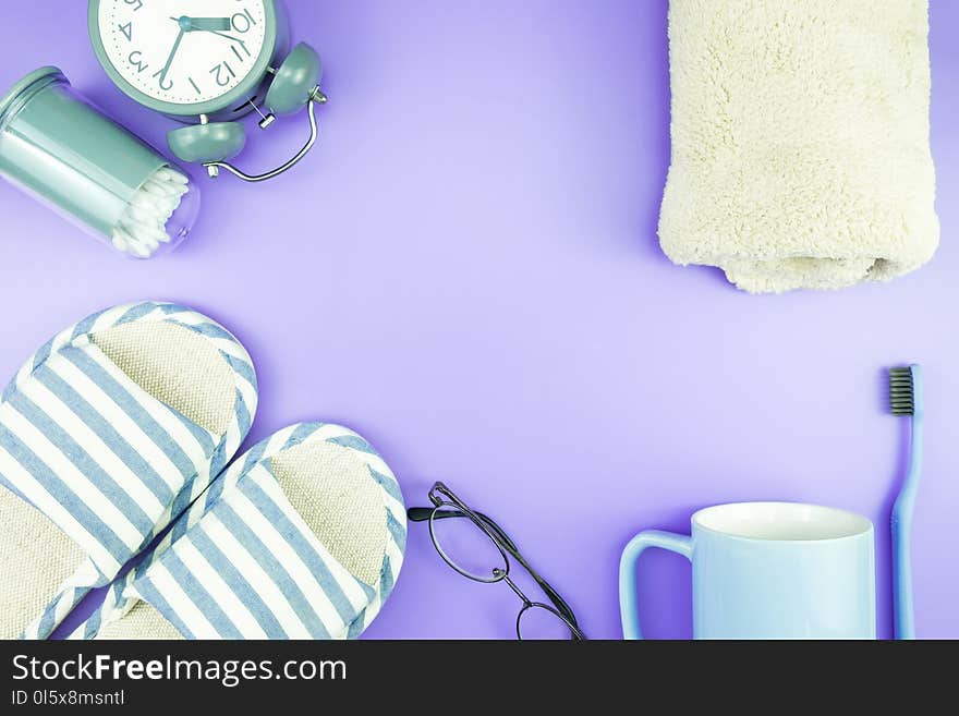 Home equipment clock flat lay composition tooth brush glasses, towel and slippers top view on blue and purple violet concept