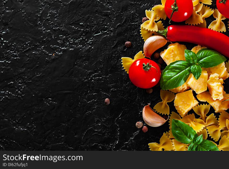 Food background. Ingredients for cooking Italian pasta - farfalle pasta, red chili peppers, cherry tomato, basil, black pepper, garlic, parmesan cheese on dark background with copy space. top view