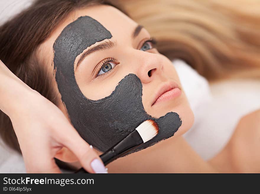 People, beauty, spa, cosmetology and skincare concept - close up of beautiful young woman lying with closed eyes and cosmetologist applying facial mask by brush in spa.