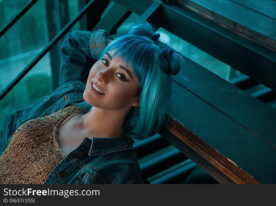 Stylish woman with a white shiny smile, wearing a blue wig, jeans and a golden blouse laying on stairs while relaxing in a loft, industrial interior. Sensual girl looking to camera.