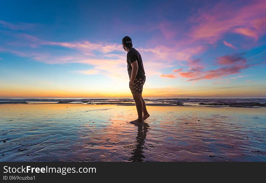 Man in Black T-shirt Wearing Cap Near Body of Water during Sunset