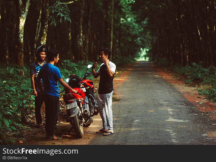 Three Men Standing Beside Red Motorcycle Surrounded With Green Trees