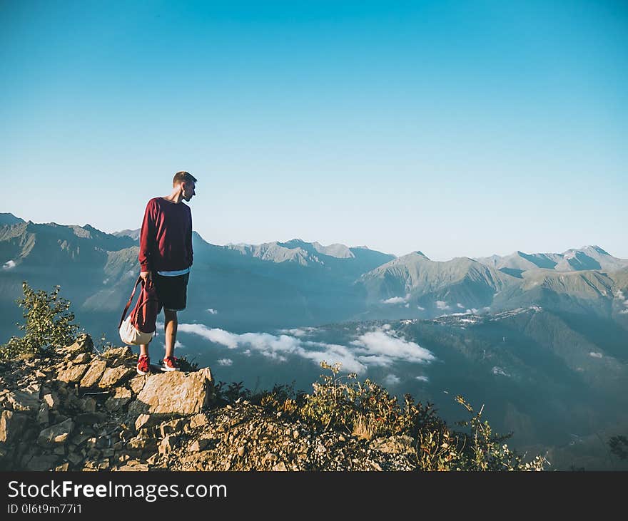 Man in Red Sweatshirt and Black Shorts Standing on Large Brown Rock on Top of a Mountain