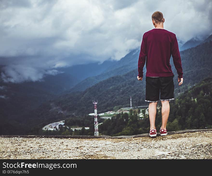 Man Wearing Maroon Sweater and Black Shorts Standing in Front of Mountain