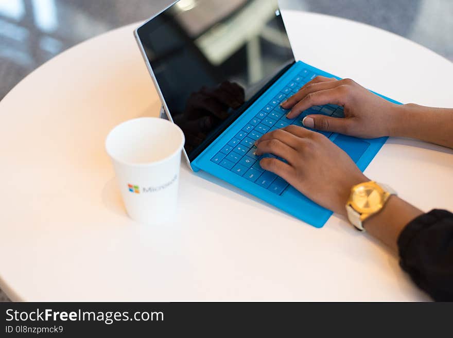 Person Wearing Round Gold-colored Watch Using Black Tablet Computer With Blue Detachable Keyboard on Round White Wooden Table