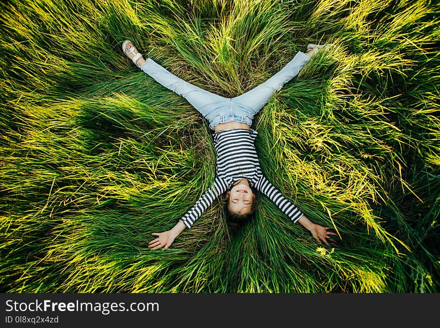 Little girl in a striped T-shirt lies on a big green glade and looks at the camera smiling. Top view. Little girl in a striped T-shirt lies on a big green glade and looks at the camera smiling. Top view.