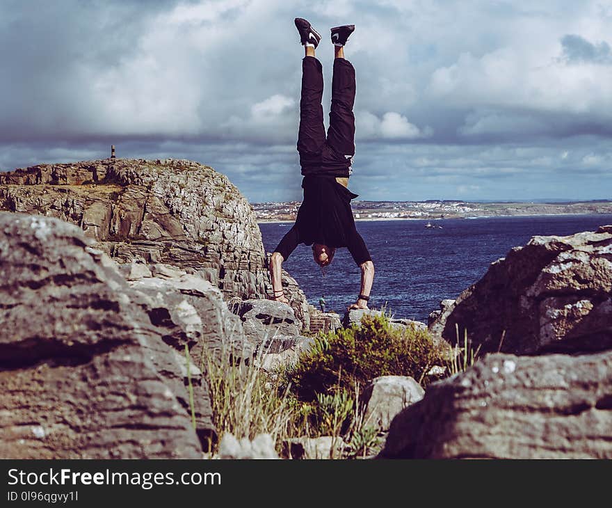 Man Wearing Black Dress Shirt and Burgundy Pants Hand Standing on Rock Formations Across Sea