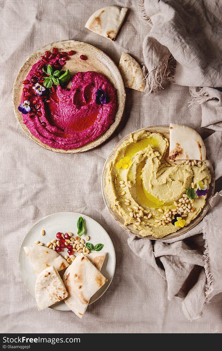 Variety of homemade traditional and beetroot spread hummus with pine nuts, olive oil, pomegranate served on ceramic plates with pita bread and green salad on linen cloth background. Flat lay, space. Variety of homemade traditional and beetroot spread hummus with pine nuts, olive oil, pomegranate served on ceramic plates with pita bread and green salad on linen cloth background. Flat lay, space.