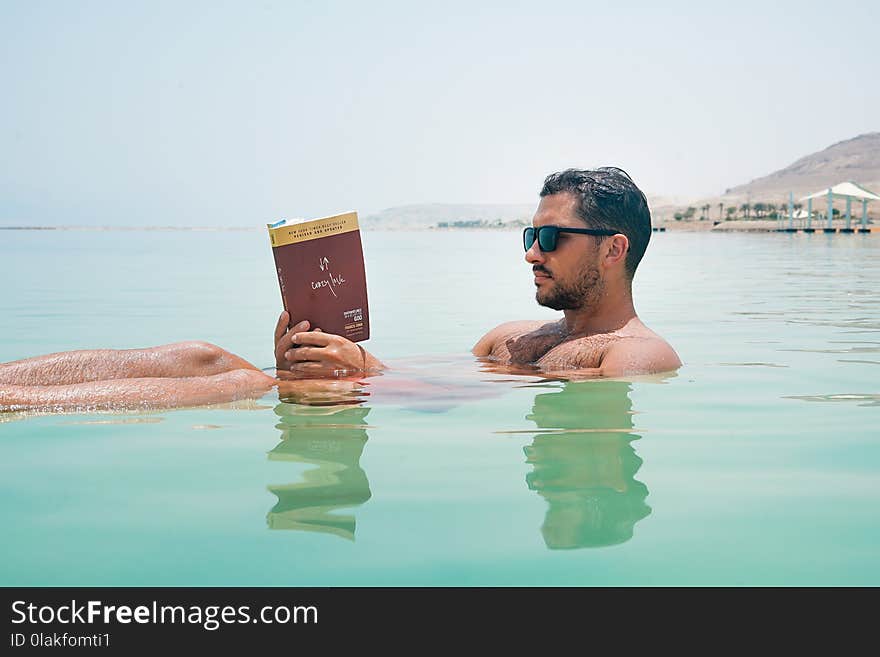 Man Wearing Sunglasses Reading Book on Body of Water