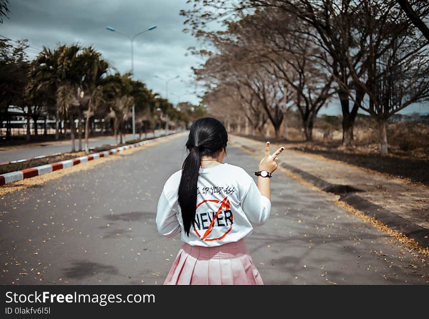 Woman Wearing White Long-sleeved Shirt and Pink Pleated Skirt Standing on Asphalt Road Between Green Trees Under Gray Sky at Daytime