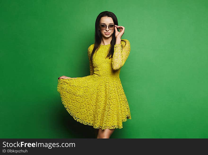 Beautiful, and fashionable brunette model girl in short yellow dress and sunglasses pulls up the hem of her dress and posing at green background at studio.