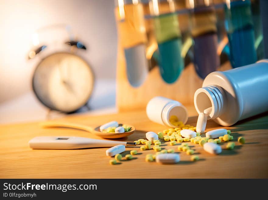 Pills spilling out of pill bottles with digital thermometer and pharmaceutical spoon on wooden board. Table clock and test tube in the background. Prescription medicine and medical treatment concepts. Pills spilling out of pill bottles with digital thermometer and pharmaceutical spoon on wooden board. Table clock and test tube in the background. Prescription medicine and medical treatment concepts