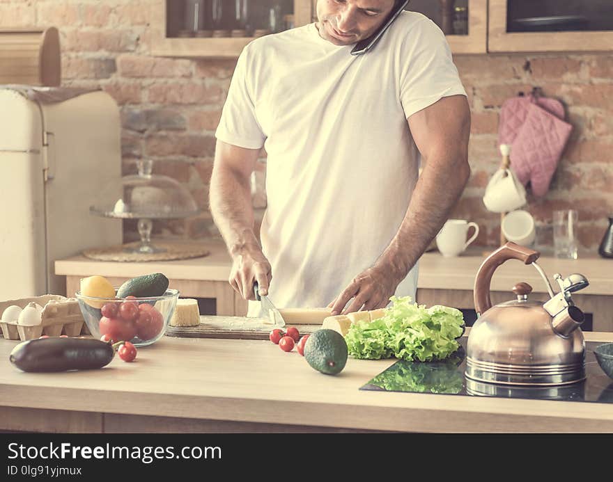 Young attractive man cutting cheese and talking on the phone in the kitchen