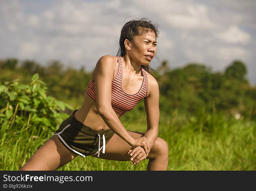 Fit and sporty middle aged runner Asian woman stretching leg and body after running workout on green field beautiful background in sport training and healthy lifestyle concept