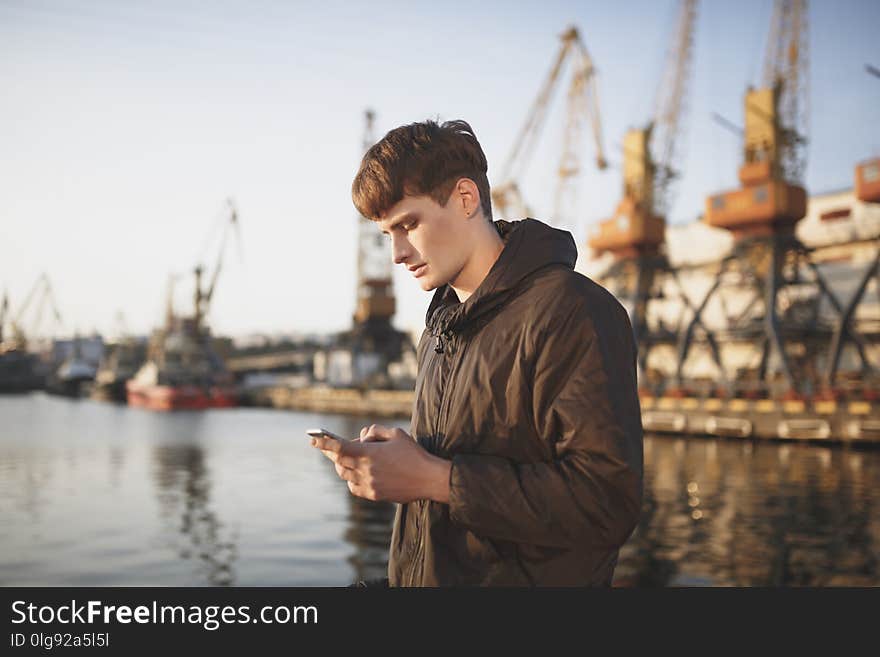 Young Man With Brown Hair Standing And Thoughtfully Looking In His Mobile Phone. Cool Boy In Down Jacket Standing With