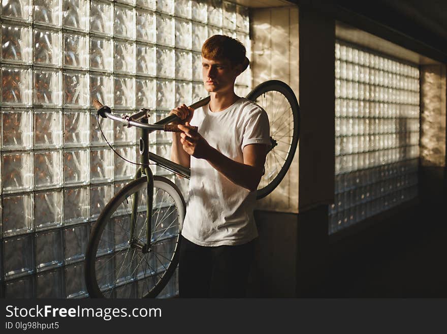 Portrait of thoughtful boy standing and holding bicycle that leaning on his shoulder while dreamily looking aside. Young man in white t-shirt standing with classic bicycle