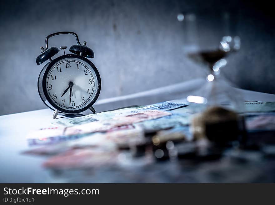 Reflection of clock and blurred hourglass on glowing table with banknotes and coins of international currency. Time investment and passing time. Urgency countdown timer for business deadline concept. Reflection of clock and blurred hourglass on glowing table with banknotes and coins of international currency. Time investment and passing time. Urgency countdown timer for business deadline concept