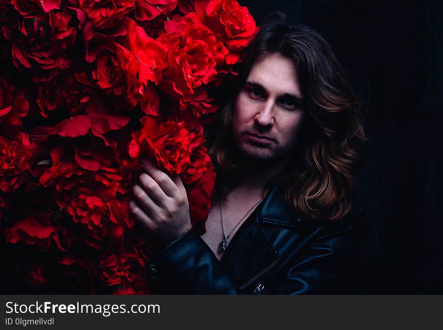 Brutal man in a leather jacket on a naked body with long hair stands near the decor with red flowers flowers. strength