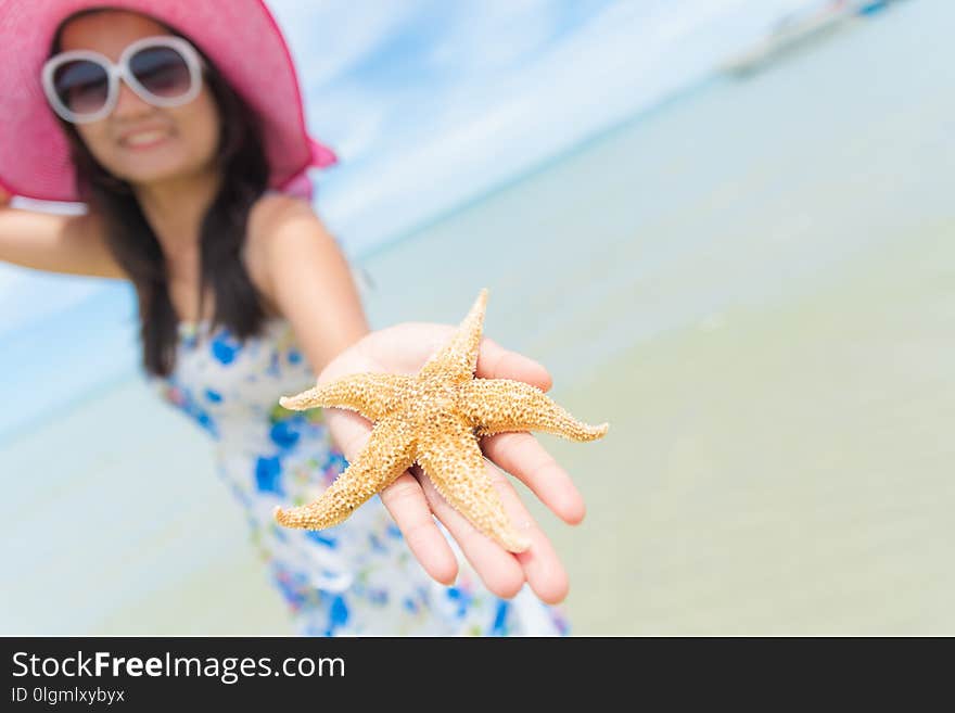 Beautiful woman wearing hat beach and sunglasses and holding starfish on hands over sandy beach, green sea and blue sky background for summer holiday and vacation concept.