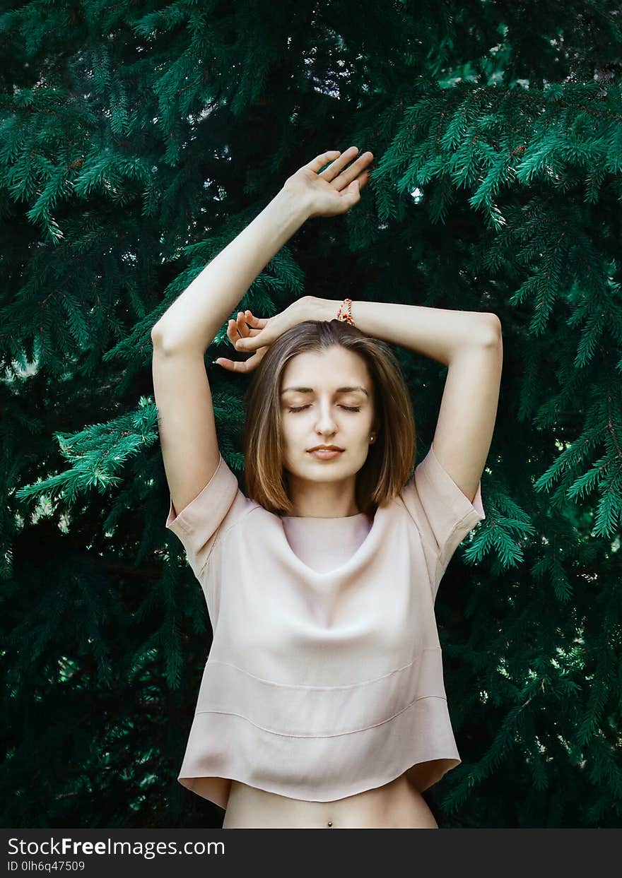 Lifestyle portrait of young white woman with short hair standing in front of pine tree with her eyes closed and hands lifted, concept of natural beauty or closeness-to-nature