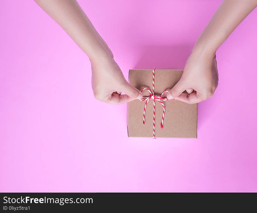 Female hands bow tie a simple craft paper gift box wrapped with colorful rope with as a present for Christmas, new year, valentine day or anniversary on pink background, top view