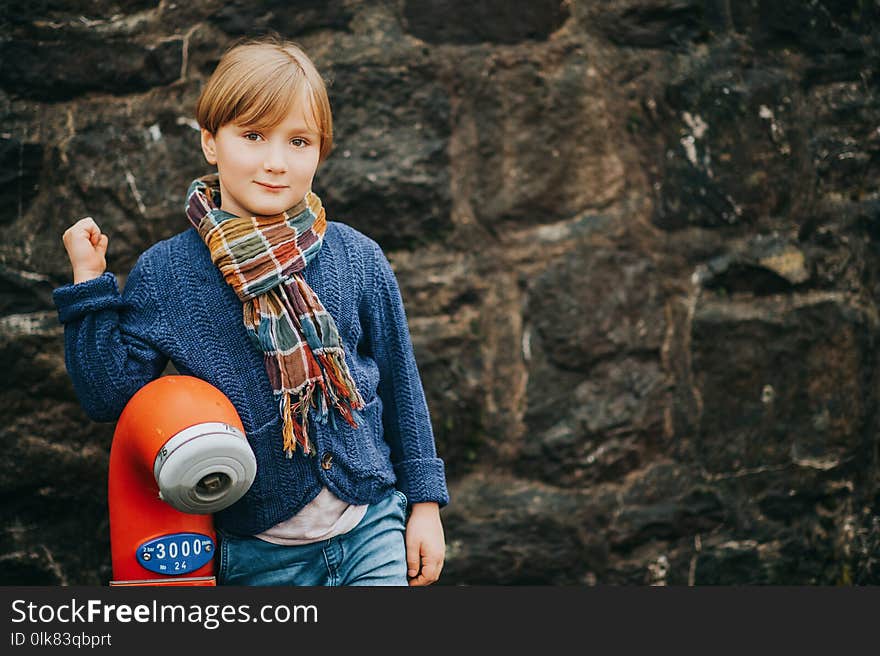 Outdoor portrait of handsome little boy wearing denim jeans, blue knitted jacket, stylish kid posing against dark wall. Fashion for small children