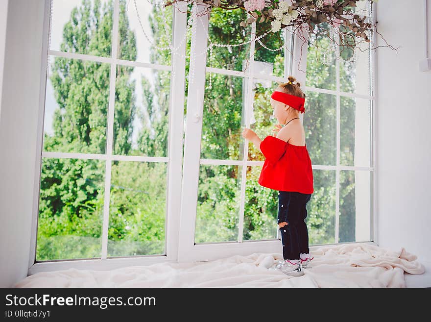 Little funny two years old baby girl in stylish red clothes and jeans and sneakers stands with back on the windowsill near the window. Headband and hairstyle on two heads in summer in sunny weather.