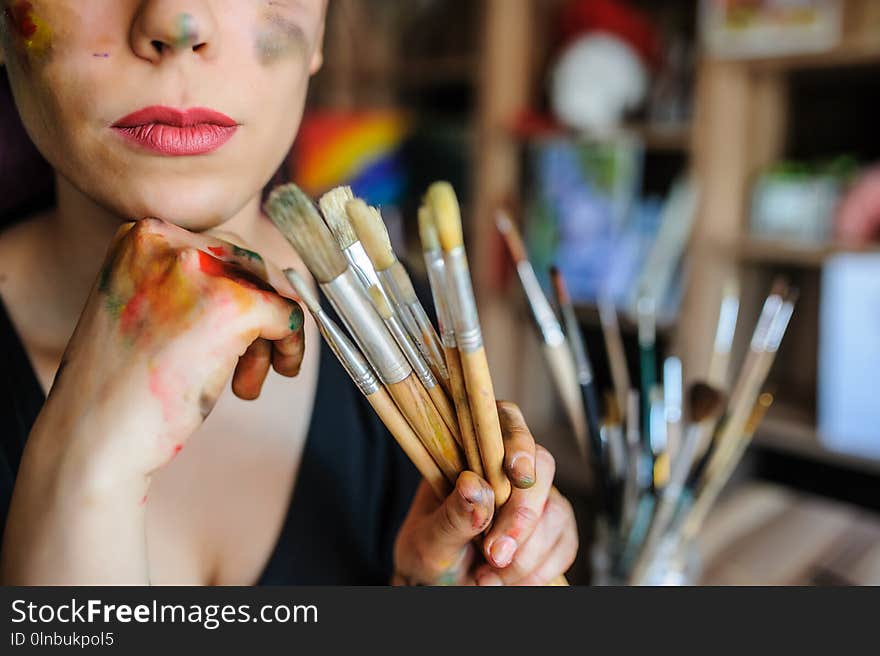 Crop picture of lips and face of beautiful female artist with purple hair and dirty hands with different paints on them, holding paintbrushes in her art studio.