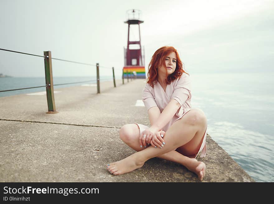 A young redheaded girl in a large round hat and pink dress sitting on the moorage near ocean. A young redheaded girl in a large round hat and pink dress sitting on the moorage near ocean