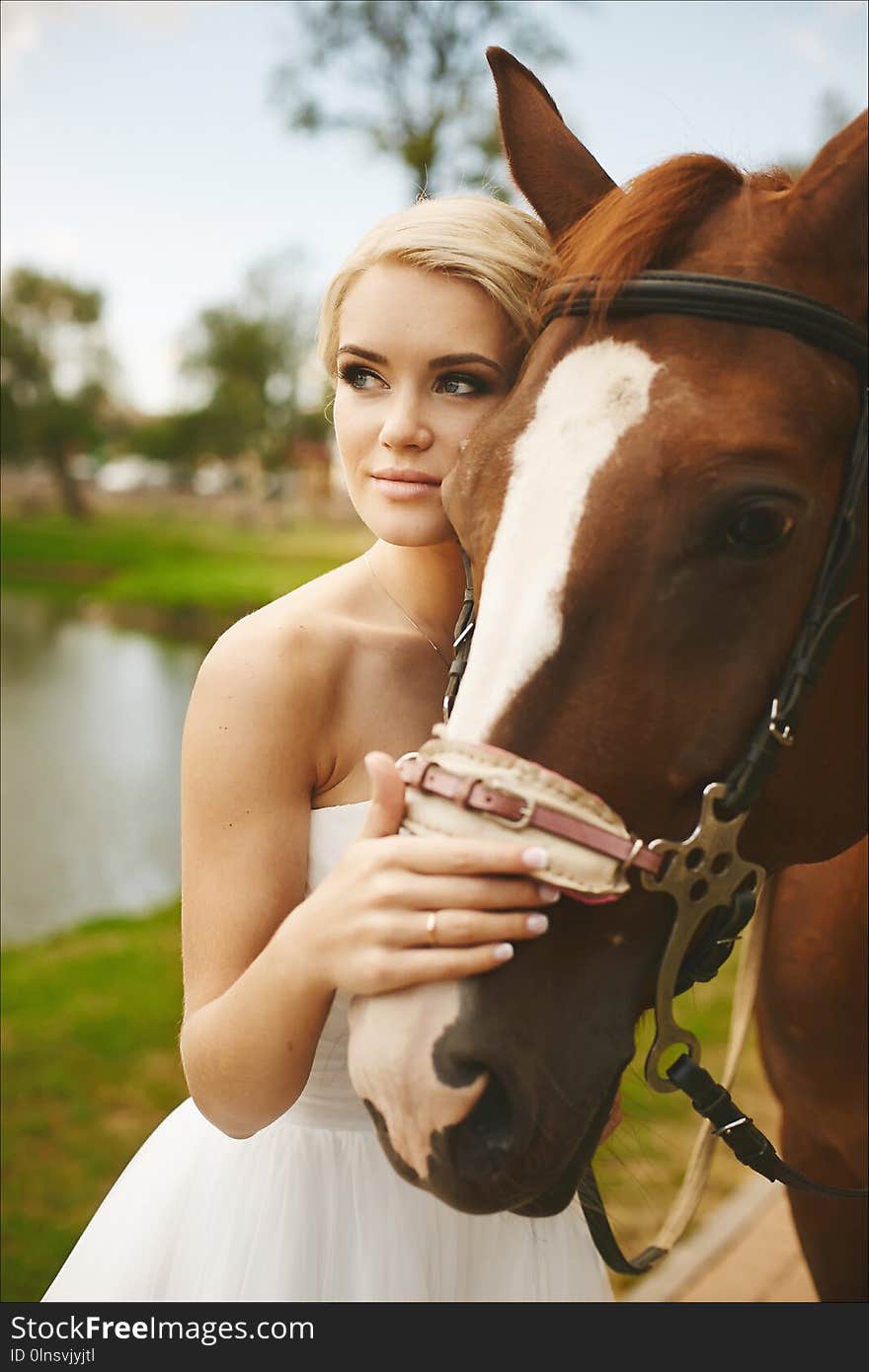 Beautiful and fashionable blonde model girl with blue eyes, with stylish hairstyle and bright makeup, in white dress posing with brown horse outdoors.