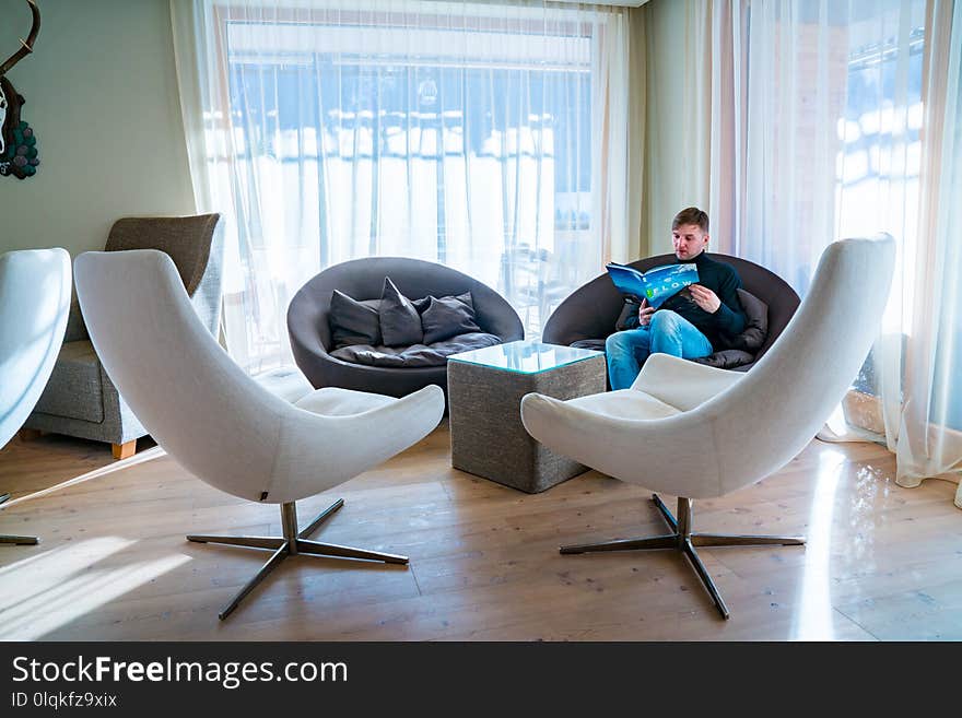 Young man sitting in a chair reading a book in a comfortable library room near huge windows.