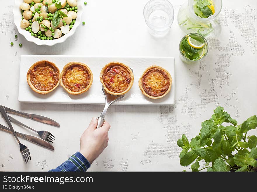 A Chef places a mini quiche on a white plate with three others on a white table with cutlery and a dish of peas and potatoes. A Chef places a mini quiche on a white plate with three others on a white table with cutlery and a dish of peas and potatoes