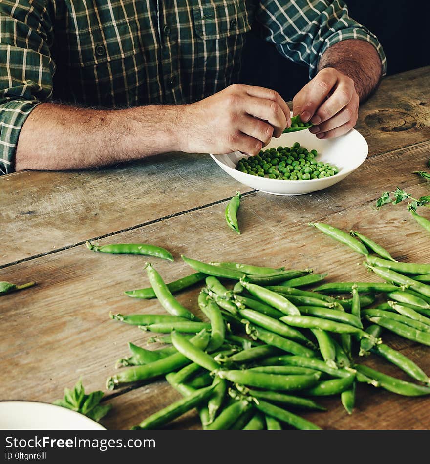 Male hands clean green peas sitting at wooden table in the kitchen. Rustic style. Green peas in bowl on wooden table. Male hands clean green peas sitting at wooden table in the kitchen. Rustic style. Green peas in bowl on wooden table