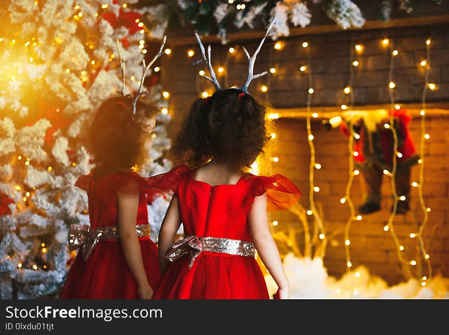 Two little girls looking at the christmas fireplace near beautiful Christmas tree. Twins in red dresses looking at Santa in fireplace. Two little girls looking at the christmas fireplace near beautiful Christmas tree. Twins in red dresses looking at Santa in fireplace.