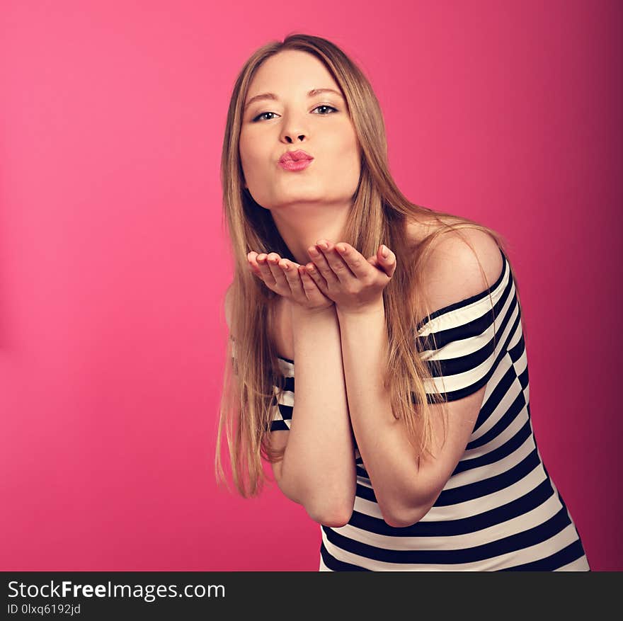 Beautiful woman with enjoying eyes showing a kissing sign and blowing the lips on the hand in fashion stripped dress on pink background. Closeup portrait