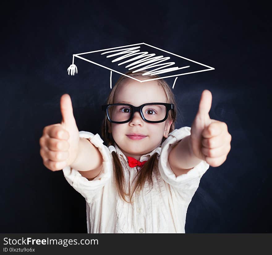 Cute schoolgirl wearing graduation hat showing thumb up in classroom. School concept