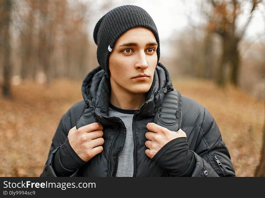 Stylish young man in a black knitted hat and jacket standing near the autumn trees.