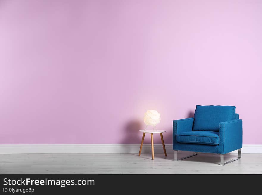 Comfortable armchair and lamp on table near color wall with space for text. Interior element