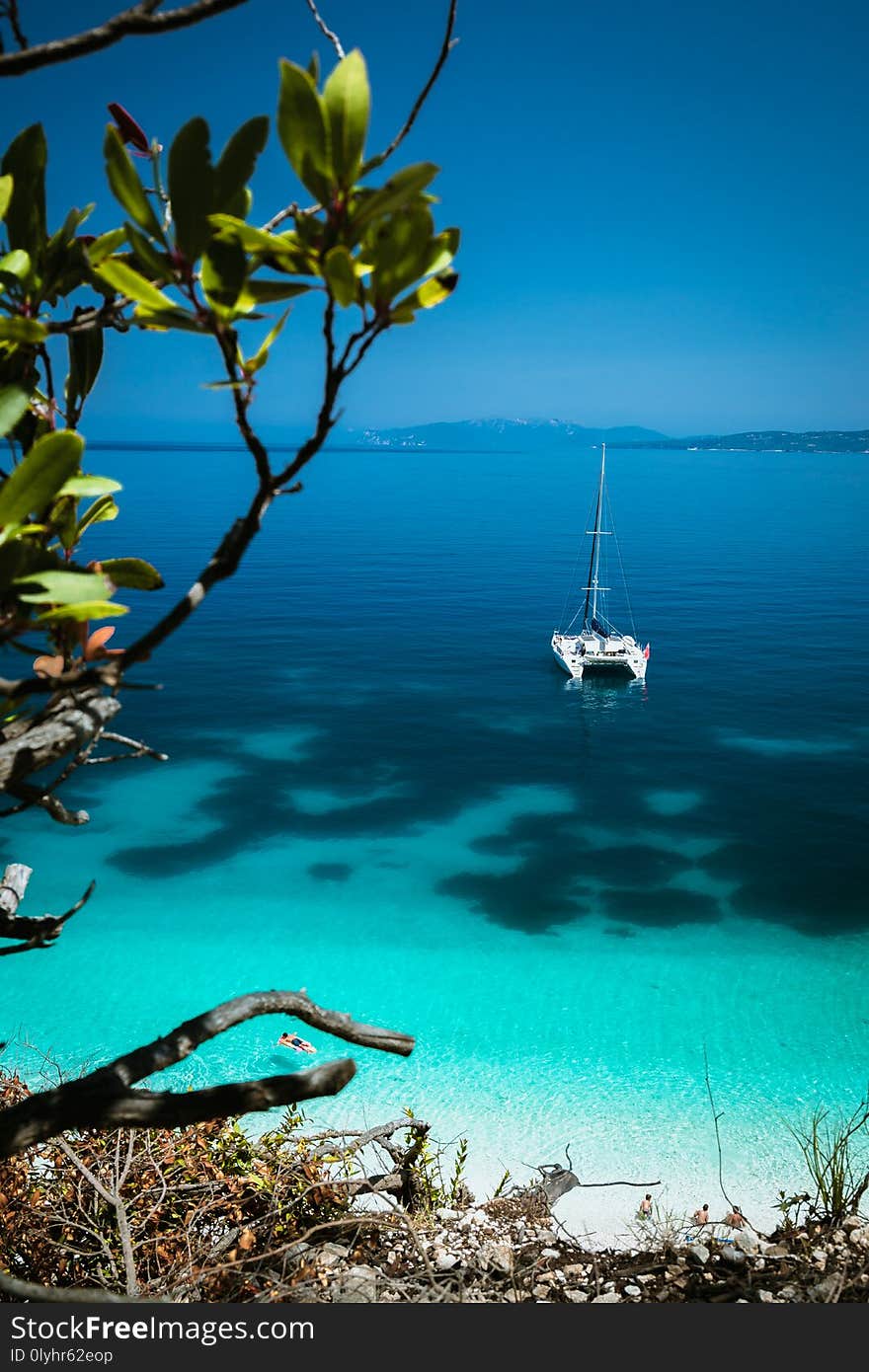 White catamaran yacht at anchor on clear azure surface with dark pattern in calm blue lagoon. Unrecognizable tourists relax on airbed in water near the beach.