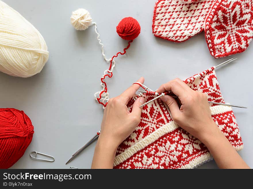 Girl knits red and white Norwegian jacquard hat knitting needles on gray wooden background. Process of knitting. Top view. Flat lay