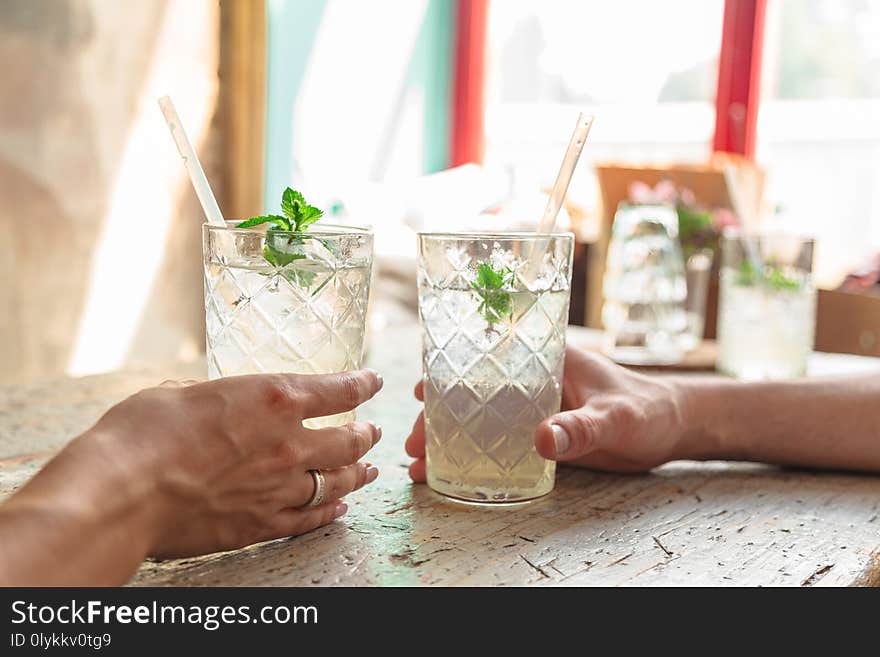 Two refreshing drinks in transparent relief glasses with white straws and mint leaves are in hands of man and woman with wedding ring on her finger in background of window and old wooden table. Two refreshing drinks in transparent relief glasses with white straws and mint leaves are in hands of man and woman with wedding ring on her finger in background of window and old wooden table.