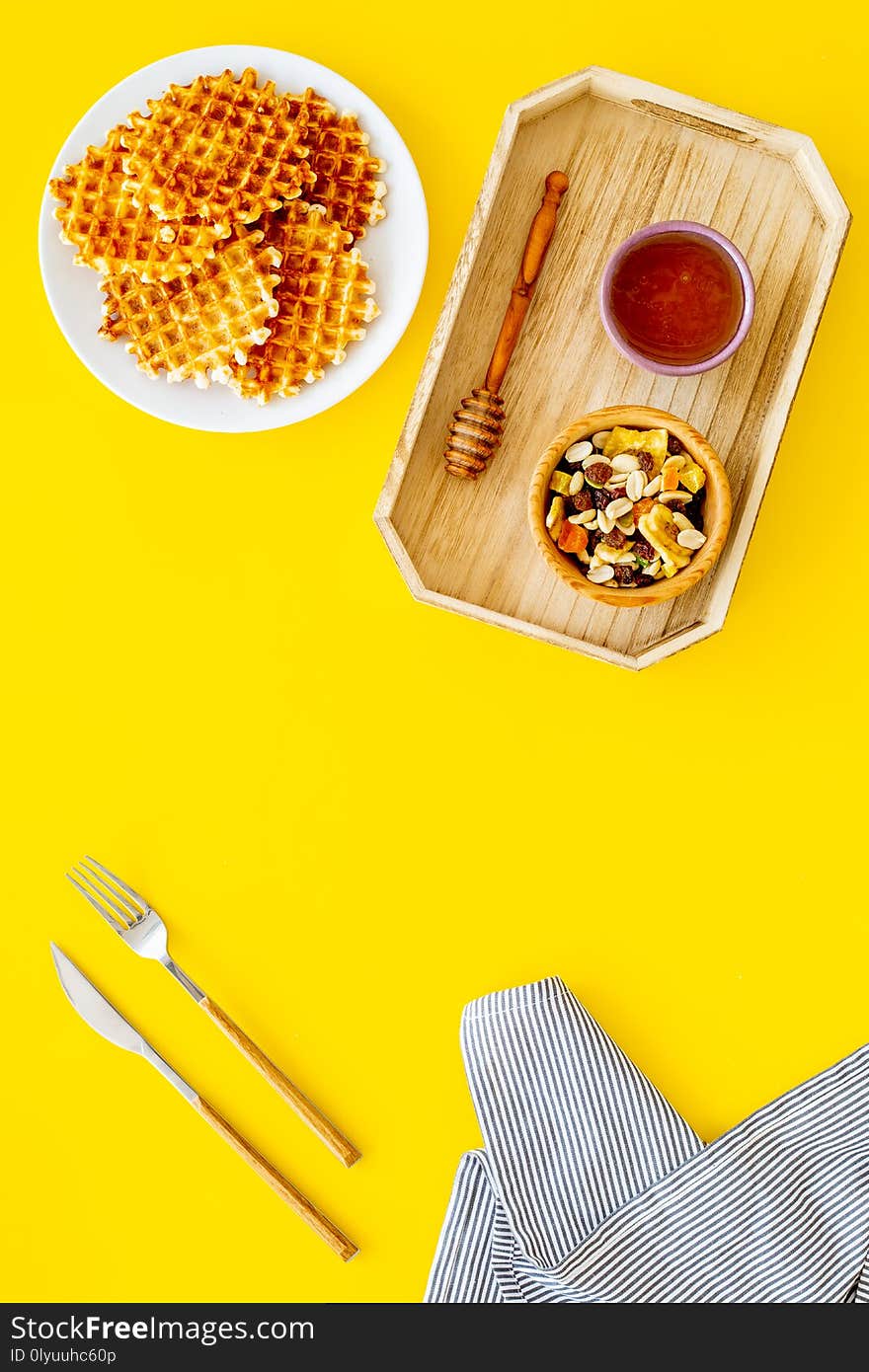 Round belgian waffles for breakfast. Breakfast in bed. Waffles on plate. Honey and dried fruits in tray, knife and fork, tablecloth on yellow background top view.