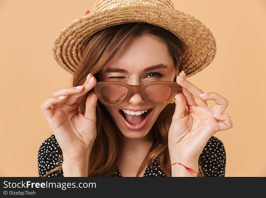 Image of summer pretty woman 20s wearing straw hat touching sunglasses and smiling at camera over beige background