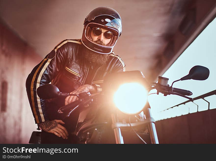 Close-up portrait of a brutal bearded biker in helmet and sunglasses dressed in a black leather jacket sitting on a retro motorcycle with included headlight.