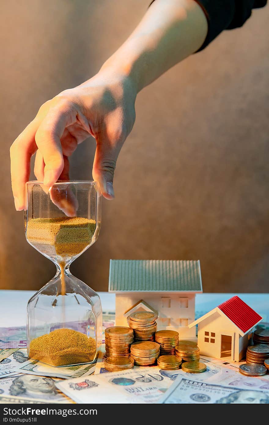 Real estate or property investment growing business. Home mortgage loan rate. Saving money for retirement concept. Male hand holding hourglass with house model and coin stack on banknotes on the table. Real estate or property investment growing business. Home mortgage loan rate. Saving money for retirement concept. Male hand holding hourglass with house model and coin stack on banknotes on the table